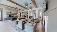 Located at 2155 Morris Basket Blvd in North Charleston, South Carolina, this single-floor amenity space is designed as a contemporary communal lounge and gathering hub. The interior is defined by soaring ceilings accented with prominent wooden beams that add warmth and architectural character to the otherwise modern, neutral palette. Light wide-plank hardwood flooring runs throughout, unifying the open-plan layout.

The main lounge area is generously proportioned, furnished with a large sectional sofa, coordinating gray armchairs, and a live-edge wood coffee table, all anchored by layered patterned area rugs. A fireplace set within a sage green board-and-batten accent wall serves as the room's focal point, topped by a wall-mounted flat-screen television. Sculptural pendant light fixtures hang overhead, adding a distinctive design element.

Adjacent to the lounge is a dining and bar station area featuring a large communal table with mid-century style seating, pendant lighting, open metal shelving, and a bold geometric-patterned wallcovering as a backdrop. Glass-paned interior partitions divide additional workspaces or private rooms beyond, while large exterior windows fill the space with natural light and views of the surrounding exterior amenities.