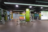 Located at 800 W Marietta St NW in Atlanta, GA, this fitness center offers a spacious, well-equipped environment designed to support a comprehensive workout experience. The facility spans a single floor with an open, industrial-style layout featuring exposed ductwork and piping against a dark ceiling, creating a modern, urban aesthetic. Durable rubber flooring covers the entire space, providing safety and comfort across all exercise zones. The cardio area is outfitted with a robust selection of TRUE-brand treadmills, ellipticals, stationary bikes, and stair climbers, positioned near large windows that allow natural light to fill the space and offer views of the surrounding greenery. The strength training zone features a wide array of cable machines, resistance equipment, and free-weight benches, complemented by floor-to-ceiling mirrors that enhance the sense of space. A dedicated open area with a folding partition wall provides flexible space for stretching, functional training, or group activity, accented by a vibrant geometric mural in pastel tones. Motivational wall text reading "CAN & WILL" adds an energizing touch to the atmosphere. Accent walls in olive green punctuate the white surroundings, contributing to the facility's cohesive and dynamic visual identity. The gym is well-lit throughout, combining recessed and linear lighting fixtures for consistent illumination across all workout zones.