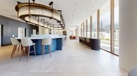Located on Bathurst Street in Toronto's vibrant King West neighbourhood, this expansive single-floor amenity space is a masterclass in contemporary design. The open-plan layout spans a generously sized floor plate, creating a fluid and versatile environment that seamlessly transitions between distinct zones.

At the heart of the space sits a striking circular bar with a bold blue base and a thick marble countertop, encircled by woven high-back bar stools with warm walnut legs. A sculptural tiered chandelier with brass and glass detailing crowns the bar, serving as a dramatic focal point. Adjacent to it, a curved dark-finished secondary bar offers additional seating along the window line.

Floor-to-ceiling glazing wraps much of the perimeter, flooding the interior with natural light and providing unobstructed views of the Toronto skyline. A dedicated dining area features a long communal wood table beneath a slatted timber ceiling with pendant globe lights, complemented by curved banquette seating upholstered in rich leather tones against a textured stone-effect feature wall.

A lounge zone with plush armchairs, a round coffee table, and a boldly patterned area rug opens toward the outdoor terrace through glass partitions. The entry area is finished with large-format stone tile flooring and curated artwork on clean white walls, lending a gallery-like refinement throughout.