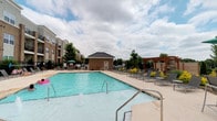The pool area offers a generously sized outdoor retreat designed for relaxation and recreation. At its center sits a large, sparkling swimming pool with crystal-clear blue water, accessible via stainless steel entry rails and steps with depth markings along the edge. The surrounding pool deck is finished in a light-toned surface that provides ample space for lounging.

A row of gray chaise lounge chairs lines one side of the deck, accented by ornamental yellow plantings that add a touch of color. A traditional wood pergola anchors one end of the space, offering a shaded gathering spot. On the opposite side, a classic brick pool house with a dark metal hip roof serves as a functional support structure for the area.

Additional seating with a market umbrella is tucked into a corner, providing a casual outdoor dining option. The entire area is enclosed by a perimeter fence, and mature trees provide natural backdrop greenery. The community buildings are visible nearby, reinforcing the sense of a cohesive, amenity-rich environment open to an expansive sky above.