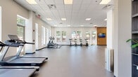 This well-appointed fitness center offers a generously sized, single-floor layout designed to accommodate a full spectrum of workout needs. The open floor plan is finished with durable speckled rubber flooring throughout, providing both safety and comfort during exercise. Large windows line multiple walls, flooding the space with natural light and offering views of the surrounding greenery, creating an energizing atmosphere for workouts.

The cardio zone features a row of treadmills positioned along windowed walls, along with elliptical trainers and stationary bikes, all arranged to maximize space and airflow. A dedicated strength training area includes a multi-station cable machine, adjustable weight benches, and free weights, supported by full-length mirrors that enhance the sense of openness. Structural columns are integrated seamlessly into the layout without impeding the generous floor space available for stretching or functional training.

A standout design element is the bold, typographic motivational mural that spans an entire accent wall, incorporating fitness-themed words and phrases that add visual energy to the environment. Warm wood-paneled wall accents and a wall-mounted flat-screen TV provide a modern, polished aesthetic. The drop ceiling with recessed panel lighting ensures consistent, even illumination throughout. Multiple entry and exit points connect the space to the outdoors, reinforcing its bright, accessible design.