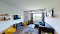This single-floor unit offers a thoughtfully organized layout encompassing a living room, two bedrooms, two full bathrooms, and a kitchen. The living room serves as the central gathering space, featuring wall-to-wall carpeting, a wall-mounted flat-screen TV, a media console, and a comfortable sectional sofa. A large window frames lush, tree-lined views, flooding the room with natural light. Bright accent seating adds a vibrant, modern touch to the space.

Both bedrooms are carpeted and share similarly panoramic views of the surrounding greenery through generously sized windows dressed with grayish-blue curtains. Each bedroom includes a dedicated closet with built-in shelving for organized storage, and ample space for a full bed and desk setup.

The two full bathrooms are identically configured, each featuring a single-sink vanity with warm wood cabinetry, a tub-shower combination, tile-style flooring, and open wire shelving for additional storage.

The galley-style kitchen is finished with warm wood cabinetry, light countertops, a double-basin stainless steel sink, and hardwood flooring. The compact, efficient layout maximizes functionality within the available space. Overall, the unit presents a clean, modern aesthetic with an emphasis on natural light and outdoor connectivity throughout.