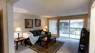 This single-floor space centers around a welcoming living room designed for both comfort and functionality. The room features a textured ceiling that adds subtle visual interest overhead, while warm, neutral-toned walls create an inviting atmosphere throughout. Carpet flooring provides a soft underfoot feel, complemented by a decorative area rug that anchors the seating arrangement.

A large sliding glass door with vertical blinds serves as a standout architectural element, flooding the room with natural light and offering a direct view of a private outdoor balcony. Glimpses of surrounding greenery and an exterior structure are visible through the glass, lending a connection to the outdoors.

The living area is furnished with a plush, dark upholstered sofa accented with decorative pillows and a draped throw blanket, paired with a classic wooden coffee table. A pair of framed wall art pieces adds personality to the space, while a traditional table lamp provides warm ambient lighting. The overall ambiance is relaxed and functional, making it a comfortable everyday living environment.