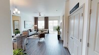 This single-floor space showcases a well-appointed, open-concept layout designed for comfortable living. The main living area is anchored by dark hardwood-style flooring that contrasts elegantly with the crisp white walls, creating a clean and modern aesthetic throughout. A ceiling fan overhead provides both function and a finishing touch to the room's design.

Natural light fills the space generously through large windows dressed with layered brown patterned drapes, complemented by a sliding glass door that opens to an outdoor area, offering views of lush greenery beyond. This seamless indoor-outdoor connection enhances the sense of openness and brightness in the room.

The living area is thoughtfully arranged with a neutral-toned sofa, accent chairs in warm brown tones, and a soft area rug that grounds the seating arrangement. Tasteful décor, including framed wall art and a tall potted plant, adds character without overwhelming the space.

A dedicated laundry area is accessible through a set of white paneled doors, neatly tucked to one side. Overall, the space exudes a calm, contemporary ambiance with a neutral palette, making it both inviting and versatile.