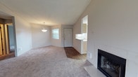 This single-floor space showcases an open-concept layout that seamlessly connects the living area, dining space, and kitchen. A standout architectural feature is the vaulted ceiling, which rises dramatically over the living room, lending a sense of openness and volume to the interior. The living area is finished with plush wall-to-wall carpet in a neutral tone, providing a warm and comfortable feel underfoot.

A tiled fireplace anchors one corner of the living room, featuring a classic white tile surround and a glass-front firebox, serving as a functional and visual focal point. Adjacent to the fireplace, the flooring transitions to hardwood-style planks near the entryway, where a paneled front door provides access to the space. Natural light filters in through a window near the dining area, which is highlighted by a simple pendant light fixture overhead.

A pass-through opening connects the living and dining areas to the kitchen, which features light wood cabinetry and a countertop visible from the main living space. The neutral color palette throughout — soft whites and warm grays — creates a cohesive, move-in ready environment. Textured ceilings complete the aesthetic of this well-proportioned, single-level home.