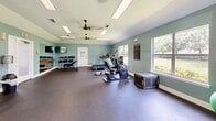 This fitness center offers a generously sized, single-room workout facility designed to accommodate a full range of exercise needs. The space is finished with soft teal-painted walls, a textured ceiling, and durable rubber flooring throughout, creating a clean and functional environment suited for regular use.

Natural light flows in through multiple windows along the perimeter walls, providing views of mature trees and greenery outside. Dual ceiling fans help maintain comfortable air circulation throughout the space, complemented by recessed fluorescent lighting for consistent illumination at any hour.

The facility is comprehensively equipped for both cardio and strength training. Cardio options include treadmills, an elliptical trainer, and a recumbent exercise bike. The strength training area features a cable machine, a power rack with barbells and weight plates, adjustable benches, and a full dumbbell rack with an accompanying mirror and wall-mounted television. Stability balls and additional free weights round out the available equipment.

A large wall mirror expands the sense of space and supports proper form during workouts. The open layout ensures ample floor space for stretching, bodyweight exercises, or functional training movements, making this fitness center a versatile and inviting space for users of all fitness levels.