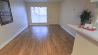 This single-floor space offers a clean, functional layout with a welcoming and understated aesthetic. The main living area is immediately notable for its warm hardwood-style flooring, which runs throughout and adds a sense of continuity and warmth to the interior. Neutral, light-toned walls keep the space feeling open and adaptable to a variety of furnishing styles. A large front-facing window floods the living area with natural light, creating a bright and airy atmosphere while offering a view of the exterior surroundings. The front entry door is positioned adjacent to this window, providing a straightforward and accessible entryway into the home. White baseboards frame the perimeter of the room, adding a crisp, finished detail to the overall design. A partial staircase railing is visible at the edge of the frame, suggesting a multi-level element or nearby stairwell access. The overall ambiance of the space is simple and well-maintained, offering a neutral canvas that is move-in ready. The combination of natural light, clean flooring, and minimalist finishes makes this a practical and comfortable living environment suited to a range of lifestyles.