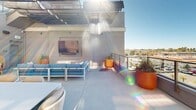 Located at 18750 Delaware St in Huntington Beach, California, this rooftop terrace is a well-appointed outdoor amenity space designed for both relaxation and social gatherings. The open-air deck is bathed in abundant Southern California sunshine, with retractable fabric awnings providing adjustable shade across multiple seating zones.

The terrace is organized into distinct functional areas. A lounge zone features a contemporary blue outdoor sofa and woven rattan accent chairs, anchored by a wall-mounted flat-screen display and complemented by vibrant orange ceramic planters with lush greenery. Separate dining areas offer white rectangular tables paired with woven rope-back chairs and wooden side chairs in warm tones, accommodating groups of varying sizes.

Large glass-paneled windows and doors connect the rooftop to an interior space, allowing natural light to flow between the two environments. A staircase with glass railings provides access between levels, reinforcing the modern architectural design. The perimeter of the deck is enclosed by sleek glass balustrades, preserving unobstructed panoramic views of the surrounding Huntington Beach cityscape and the clear blue sky beyond. The clean concrete flooring, crisp white structural walls, and contemporary furnishings give the space a cohesive, resort-inspired aesthetic ideal for outdoor entertaining.