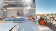 Located at 18750 Delaware St in Huntington Beach, California, this rooftop terrace is a well-appointed outdoor amenity space designed for both relaxation and social gatherings. The open-air deck is bathed in abundant Southern California sunshine, with retractable fabric awnings providing adjustable shade across multiple seating zones.

The terrace is organized into distinct functional areas. A lounge zone features a contemporary blue outdoor sofa and woven rattan accent chairs, anchored by a wall-mounted flat-screen display and complemented by vibrant orange ceramic planters with lush greenery. Separate dining areas offer white rectangular tables paired with woven rope-back chairs and wooden side chairs in warm tones, accommodating groups of varying sizes.

Large glass-paneled windows and doors connect the rooftop to an interior space, allowing natural light to flow between the two environments. A staircase with glass railings provides access between levels, reinforcing the modern architectural design. The perimeter of the deck is enclosed by sleek glass balustrades, preserving unobstructed panoramic views of the surrounding Huntington Beach cityscape and the clear blue sky beyond. The clean concrete flooring, crisp white structural walls, and contemporary furnishings give the space a cohesive, resort-inspired aesthetic ideal for outdoor entertaining.