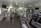 Located at 35101 Harper Ave in Clinton Township, Michigan, this single-floor fitness facility is thoughtfully designed to support a well-rounded workout experience. The main exercise area features durable rubberized flooring and is equipped with a range of cardio machines, including treadmills, an elliptical trainer, and a stationary bike, alongside a multi-station strength training machine. Large wall-mounted mirrors line the workout space, amplifying the sense of openness and allowing users to monitor their form. Motivational quotes are painted along the accent wall, adding an energizing atmosphere to the room. Recessed lighting throughout provides consistent, functional illumination. Separate men's and women's restrooms are conveniently accessible directly from the main workout area, along with a bank of lockers for storage. A connecting corridor, finished with patterned carpet tile flooring and neutral gray walls, leads further into the facility and includes a water fountain and informational wall-mounted displays. The overall design is clean and modern, with a practical, no-frills aesthetic focused on functionality and ease of use for fitness enthusiasts.
