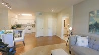 This well-appointed single-floor residence offers a thoughtfully designed open layout that promotes a sense of spaciousness and ease of movement. The living area greets you with warm hardwood-style flooring, soft neutral walls, and recessed lighting that creates a welcoming ambiance. A plush area rug anchors the seating space, which features light gray upholstered furniture complemented by a stylish floor lamp, adding both function and character to the room.

The kitchen is open to the main living space and is outfitted with white cabinetry, white appliances — including a microwave, stove, and refrigerator — and a light-toned countertop that runs along the perimeter. A breakfast bar with seating provides a casual dining option while maintaining the open flow of the space.

Two white interior doors are visible from the central hallway area — one leading to a closet and the other serving as the main entry door, both featuring classic arched panel detailing. An arched passageway leads to what appears to be a bathroom or vanity area, glimpsed with white cabinetry and warm lighting. The consistent use of white trim, neutral paint, and cohesive flooring throughout ties the entire space together in a clean, livable aesthetic.