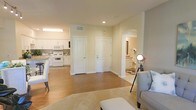 This well-appointed single-floor residence offers a thoughtfully designed open layout that promotes a sense of spaciousness and ease of movement. The living area greets you with warm hardwood-style flooring, soft neutral walls, and recessed lighting that creates a welcoming ambiance. A plush area rug anchors the seating space, which features light gray upholstered furniture complemented by a stylish floor lamp, adding both function and character to the room.

The kitchen is open to the main living space and is outfitted with white cabinetry, white appliances — including a microwave, stove, and refrigerator — and a light-toned countertop that runs along the perimeter. A breakfast bar with seating provides a casual dining option while maintaining the open flow of the space.

Two white interior doors are visible from the central hallway area — one leading to a closet and the other serving as the main entry door, both featuring classic arched panel detailing. An arched passageway leads to what appears to be a bathroom or vanity area, glimpsed with white cabinetry and warm lighting. The consistent use of white trim, neutral paint, and cohesive flooring throughout ties the entire space together in a clean, livable aesthetic.
