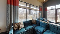 This single-floor space makes a striking first impression with its vibrant, carefully curated interior design. The main lounge area is anchored by a large teal velvet sectional sofa accented with geometric patterned throw pillows, set against dark hardwood-style flooring that extends throughout the space. A bold blue accent wall frames a sleek floating wood shelf and fireplace, adding warmth and visual depth to the room.

Floor-to-ceiling windows with black metal framing line the exterior wall, flooding the interior with abundant natural light and offering expansive views of the surrounding treetops and adjacent buildings. Dramatic patterned drapes with bold red accents frame the windows, adding a layer of color and texture. A set of glass doors opens directly onto a covered balcony, featuring stone-clad pillars and a railing that overlooks an outdoor amenity area below.

The space flows openly into a kitchen and dining area, visible through a wide architectural passage flanked by deep teal accent niches adorned with sculptural wall art and decorative spherical pieces. The kitchen features pendant lighting, a large island with bar seating, and a textured tile backsplash wall. The ceiling transitions between warm golden and neutral tones, adding dynamic visual interest throughout and reinforcing the space's bold, contemporary aesthetic.