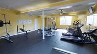 This fitness center offers a dedicated space for exercise and wellness, thoughtfully laid out across a single floor. The room is immediately recognizable by its vibrant yellow walls, which create an energizing atmosphere conducive to an active workout environment. Crown molding and white structural columns add a clean, finished look to the space.

The flooring consists of durable rubber matting throughout, providing both safety and support during exercise. A ceiling fan and recessed lighting ensure the space remains comfortable and well-lit at all times, while a window along one wall allows natural light to filter in.

The fitness area is equipped with a multi-station cable and weight machine, ideal for a variety of strength training exercises. Additional equipment includes a weight bench with free weights, a treadmill, an elliptical trainer, and a stationary bike, offering comprehensive cardio and strength options. Instructional fitness posters are displayed on the walls, providing guidance for users.

A private entrance door offers convenient and direct access to the space. Overall, this fitness center is a well-organized and functional environment that accommodates a broad range of fitness goals in a bright, motivating setting.