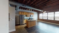 This urban loft showcases a compelling industrial aesthetic throughout its single floor, combining raw architectural elements with functional modern living. The open-concept main living and kitchen area is defined by striking exposed wood beam ceilings with visible ductwork running overhead, complemented by rich dark hardwood flooring and a ceiling fan that anchors the living space. A large multi-pane window set within an exposed brick accent wall floods the area with natural light and provides views of the surrounding cityscape, enhancing the loft's urban character.

The kitchen is well-appointed with warm wood cabinetry, granite countertops, a central island for added prep space, and a full suite of stainless steel appliances including a refrigerator, electric range, over-the-range microwave, and dishwasher. Track lighting runs along the ceiling, providing targeted illumination throughout.

A hallway leads to the private areas of the unit, featuring a carpeted bedroom with exposed wood beam ceiling details, ample closet storage, and a full bathroom complete with a tub/shower combination, single vanity with mirror, and tile flooring. The overall ambiance blends industrial rawness with residential warmth, creating a distinctive and character-rich living environment.
