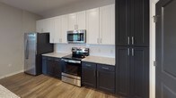 Located at 15921 White St in Charlotte, NC, this single-floor home offers a thoughtfully designed layout with contemporary finishes throughout. The kitchen is a standout feature, showcasing a striking two-tone cabinet scheme with dark lower cabinets and crisp white upper cabinetry, complemented by granite-look countertops, a classic subway tile backsplash, and a full suite of stainless steel appliances including a French door refrigerator, electric range, and built-in microwave. A peninsula with pendant lighting connects the kitchen to the open living area, which features durable hardwood-style flooring that transitions into plush carpeting in the main living space. Natural light flows through multiple windows dressed with white blinds, creating a bright and welcoming atmosphere. The bedrooms are generously proportioned, each equipped with a ceiling fan and large windows that offer outdoor views. At least one bedroom includes access to a walk-in closet with built-in shelving. The bathroom features wood-style flooring, a navy vanity with granite countertop, a tiled tub-shower combination, and a natural light-admitting window. Throughout the home, dark interior doors provide a bold contrast against the neutral warm-gray walls and white trim, lending the space a modern, cohesive aesthetic.