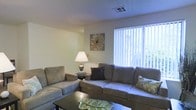 This single-floor residence offers a warm and inviting atmosphere designed for comfortable everyday living. The living room serves as the heart of the home, showcasing a neutral color palette of soft whites and warm beige tones that create a calm, understated ambiance. A large window fitted with vertical blinds allows generous natural light to fill the space, offering a bright and airy feel while providing a view of the lush greenery outside. The living area is thoughtfully arranged with a plush sectional sofa and a coordinating armchair, both upholstered in a soft taupe fabric, providing ample seating for relaxing or entertaining. A dark wood coffee table anchors the seating arrangement, adding a grounding contrast to the lighter furnishings. A classic table lamp adds a warm accent, complementing the overall cozy feel of the room. Decorative touches, including accent pillows and wall art, add subtle personality without overwhelming the clean, simple aesthetic. The ceiling features a standard textured finish with a central HVAC vent, reflecting a practical and well-maintained interior. Overall, this space presents a straightforward, comfortable layout with an emphasis on natural light and easy livability.