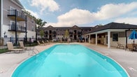 Located at 882 Taylor Station Rd in Columbus, Ohio, this residential community amenity space offers a well-appointed outdoor environment designed for relaxation and recreation. The centerpiece of the space is a generously sized swimming pool with clear blue water, surrounded by a wide concrete deck that provides ample room for lounging. Several reclining chairs are positioned around the pool area, offering residents comfortable spots to unwind under open skies.

Adjacent to the pool is a covered patio structure, providing a shaded gathering space ideal for outdoor entertaining or casual seating. The patio area is equipped with additional seating, making it a versatile spot for both quiet relaxation and social gatherings. The surrounding grounds are landscaped with young trees that add a touch of greenery to the environment.

The backdrop of the amenity area is framed by a multi-story residential building featuring a classic architectural design with dark neutral siding, gabled rooflines, and private balconies. A black iron perimeter fence defines the space, providing both security and a clean visual boundary. The overall ambiance is welcoming and community-oriented, offering residents a practical and attractive outdoor retreat within the Columbus area.