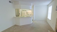 This single-floor residence is designed with an open-concept layout that seamlessly connects the kitchen and living area, creating a cohesive and functional living environment. The kitchen is outfitted with white cabinetry that extends to the upper portion of the walls, maximizing storage space. White appliances, including a refrigerator and range, are neatly integrated into the design. A peninsula-style counter with a breakfast bar serves as a natural divider between the kitchen and the adjoining living space, offering both additional prep space and casual seating. Under-cabinet lighting provides focused illumination over the work surfaces. The living area features neutral wall-to-wall carpeting that adds warmth and softness underfoot. Throughout the space, white walls and a textured white ceiling contribute to a clean, bright atmosphere. The open sightlines between the kitchen and living room enhance the sense of spaciousness, making the layout feel airy and connected. The overall aesthetic is light and minimal, offering a versatile canvas well-suited for a variety of interior styles and furnishing choices.