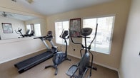This single-floor fitness space is designed to support a well-rounded cardio workout routine. The room is outfitted with three key pieces of exercise equipment: a treadmill, an upright stationary bike, and an elliptical cross-trainer, providing users with a variety of low-impact and moderate-intensity cardio options. A large wall-mounted mirror on one side of the room enhances the sense of space while allowing users to monitor their form during exercise. Natural light flows in through multiple windows fitted with horizontal blinds, creating a bright and airy atmosphere that contributes to an energizing workout environment. The walls are finished in a neutral, warm beige tone that keeps the space feeling clean and welcoming. A fitness reference poster is displayed on the wall, offering workout guidance to users. The flooring features a durable, textured carpet suited for a fitness setting. The overall layout is practical and efficiently organized, making good use of the available floor space while keeping pathways accessible between machines.