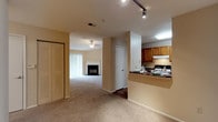 This single-floor residential unit offers a thoughtfully designed open-concept layout that seamlessly connects the main living areas. The living room is a welcoming centerpiece, anchored by a classic fireplace and fitted with plush neutral-toned carpeting that extends throughout the space. A ceiling fan provides both functional comfort and a finishing touch to the room's relaxed ambiance. Natural light filters in through large window treatments, lending the space a bright and airy feel.

The kitchen is positioned adjacent to the living area and features warm wood cabinetry, dark granite-style countertops, and a raised breakfast bar that serves as a natural divider between the kitchen and the rest of the open floor plan. A range with overhead ventilation and a skylight-style ceiling fixture add both practicality and brightness to the culinary space. Track lighting in the transitional hallway area provides additional illumination throughout.

A spacious closet with bifold doors offers generous storage, while an interior door leads to additional private spaces within the unit. The neutral color palette of beige and warm tones throughout creates a cohesive and inviting atmosphere, making this a comfortable and functional single-level living space.