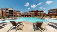 Located in Bentonville, Arkansas, this single-story community clubhouse serves as the social and administrative hub of a multi-building residential community. The exterior showcases classic brick architecture set against a expansive resort-style pool, complete with a covered pavilion, fountain features, and generous sun deck furnished with lounge chairs — ideal for outdoor relaxation.

The interior leasing office greets visitors with rich hardwood flooring, coffered ceilings with dark exposed beams, recessed lighting, and elegantly appointed workstations paired with teal patterned accent chairs. A bold blue accent wall and curated artwork add character throughout the communal lounge, which features a large sectional seating arrangement, flat-screen television, and decorative fireplace console.

A fully equipped kitchen area offers dark cabinetry, granite countertops, stainless steel appliances, and a double sink — well-suited for resident gatherings. An adjacent dining area with floor-to-ceiling windows overlooks a children's playground and the broader community grounds, filling the space with natural light.

A dedicated business center provides granite-topped workstations with computers, positioned alongside interior windows that offer direct views into the fitness facility, which is equipped with cardio machines and strength-training equipment.