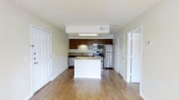 This single-floor residence offers a clean, functional layout centered around an open-concept kitchen and living area. The kitchen is a standout feature, equipped with stainless steel appliances including a refrigerator, range, and built-in microwave. Rich dark wood cabinetry provides ample storage, while granite countertops add a touch of elegance. A central kitchen island with a light-colored base offers additional prep space and serves as a natural divider between the kitchen and the open living area. The space is finished with warm-toned hardwood flooring that runs throughout, contributing to a bright and cohesive feel. Overhead, a recessed skylight panel above the kitchen enhances the sense of openness and brings in supplemental light. The neutral wall tones and white trim create a clean, versatile backdrop throughout. Multiple doorways visible from the main living area suggest well-organized access to additional rooms, offering a practical and efficient floor plan. The overall ambiance is modern yet inviting, combining durable finishes with thoughtful design elements that make the space both functional and visually appealing.