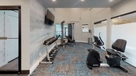 Located at 6236 Twin Oaks Dr in Colorado Springs, Colorado, this single-floor fitness center is designed to provide a comprehensive workout environment with a clean, modern aesthetic. The space is finished with stylish patchwork carpet tiles in muted blue and gray tones, recessed ceiling lighting, and crisp white walls that create a bright and energizing atmosphere.

A standout feature is the bold motivational typography mural that spans one full wall, displaying words such as "Strength," "Achieve," "Balance," "Inspire," and "Wellness," reinforcing the space's focus on health and well-being. A large flat-screen television is mounted above the mural, offering entertainment during workouts.

The cardio area is well-equipped with elliptical trainers, an upright stationary bike, and a rowing machine, complemented by a large wall mirror that enhances the sense of space. On the strength-training side, the facility includes a multi-function cable machine, an adjustable weight bench, and a full rack of dumbbells spanning a wide range of weights.

A frosted-glass sliding door provides access to an adjacent room, adding a modern architectural touch while maintaining an open and airy feel. The thoughtful layout ensures ample room to move between equipment, making this fitness center a well-rounded facility suited for a variety of workout routines.