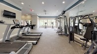 This spacious fitness center is thoughtfully designed to support a comprehensive workout experience within a single, open-plan floor. The interior is finished with neutral beige walls accented by a distinctive teal crown molding band, recessed lighting, and ceiling fans that contribute to a clean, well-ventilated atmosphere. Durable, textured carpeted flooring spans throughout, providing comfort underfoot across all exercise zones.

The cardio section is generously stocked with a row of TRUE treadmills, elliptical trainers, upright and recumbent stationary bikes, and stair climbers, all arranged to maximize space and flow. A cable crossover strength machine and additional resistance equipment occupy a dedicated strength training area, complemented by a full dumbbell rack and a medicine ball tower offering a range of weighted options. Large mirrors line portions of the walls, enhancing the sense of space and allowing users to monitor their form.

Natural light filters through multiple large windows along one end of the room, offering views of surrounding greenery. Wall-mounted televisions are positioned throughout for entertainment. Motivational wall decals add an encouraging, energetic ambiance. A small refrigerator and waste receptacle provide practical convenience. The overall design balances functionality with an inviting, polished aesthetic.