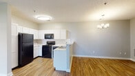 Located at 6700 E Virginia St in Evansville, Indiana, this single-story unit offers a clean, contemporary design characterized by consistent wood-look flooring and neutral gray walls throughout. The open-concept layout connects the kitchen and living area seamlessly, creating a spacious and functional main living space. The kitchen is well-appointed with white shaker-style cabinetry, stone-look countertops, a subway tile backsplash, and black appliances including a refrigerator, range, and built-in microwave. A peninsula with an integrated sink provides additional counter space and separates the kitchen from the living area, which is illuminated by a stylish chandelier. The living and dining areas benefit from access to natural light through multiple windows dressed with horizontal blinds, and a ceiling fan adds comfort to the brighter rooms. Two bathrooms are present—one featuring a tub-shower combination and another with a walk-in shower—both finished with white vanities and wood-look flooring. Bedrooms offer dedicated closet space and generous natural light, maintaining the unit's bright and airy atmosphere throughout.