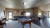 This single-floor entertainment space is thoughtfully designed for recreation and social gatherings. At its center sits a classic wooden pool table with ornate carved legs, illuminated by an industrial-style pendant light that adds a bold focal point to the room. A wall-mounted flat-screen television is positioned for easy viewing from multiple angles, making the space as suitable for watching sports as it is for friendly competition.

A full-length shuffleboard table lines the back wall, accompanied by a foosball table, ensuring a variety of gaming options for guests. One of the most striking architectural features is the floor-to-ceiling stacked stone fireplace column, which adds warmth, texture, and a rustic elegance to the otherwise modern interior. The fireplace insert provides both ambiance and comfort.

Natural light filters in through multiple windows, brightening the neutral-toned walls and complementing the recessed lighting overhead. The flooring transitions between tile and carpet, subtly defining different zones within the space. With its combination of carefully chosen furnishings, standout stonework, and versatile layout, this game room delivers a compelling and inviting atmosphere for entertainment of all kinds.