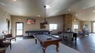 This single-floor entertainment space is thoughtfully designed for recreation and social gatherings. At its center sits a classic wooden pool table with ornate carved legs, illuminated by an industrial-style pendant light that adds a bold focal point to the room. A wall-mounted flat-screen television is positioned for easy viewing from multiple angles, making the space as suitable for watching sports as it is for friendly competition.

A full-length shuffleboard table lines the back wall, accompanied by a foosball table, ensuring a variety of gaming options for guests. One of the most striking architectural features is the floor-to-ceiling stacked stone fireplace column, which adds warmth, texture, and a rustic elegance to the otherwise modern interior. The fireplace insert provides both ambiance and comfort.

Natural light filters in through multiple windows, brightening the neutral-toned walls and complementing the recessed lighting overhead. The flooring transitions between tile and carpet, subtly defining different zones within the space. With its combination of carefully chosen furnishings, standout stonework, and versatile layout, this game room delivers a compelling and inviting atmosphere for entertainment of all kinds.