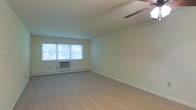 This single-floor space features a spacious, open living area finished with wall-to-wall carpeting in a neutral tone, lending a clean and understated aesthetic throughout. The room is bathed in natural light through a large multi-pane window that overlooks lush greenery, creating a bright and airy atmosphere. A ceiling fan is centrally mounted overhead, providing both ventilation and a subtle decorative element. Climate control is supported by a wall-mounted window air conditioning unit positioned beneath the window, complemented by baseboard heating that runs along the perimeter of the room — offering year-round comfort. The walls are finished in a soft, light tone that enhances the sense of openness, while white baseboards provide a clean contrast against the carpeted floor. The layout offers generous open floor space, making it highly versatile for a variety of furniture arrangements. Overall, the space presents a functional and well-lit environment with a simple, comfortable character suited for everyday living.