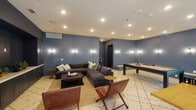 This single-floor amenity space is designed as a multifunctional social hub, combining comfortable lounging with active recreation. The interior is unified by a distinctive slate-blue wall treatment accented by elegant wall sconces, which provide warm, ambient lighting throughout. Recessed ceiling lights supplement the sconces, ensuring the space is well-lit across all activity zones.

The lounge area anchors one side of the room, centered around a generously sized sectional sofa in deep charcoal upholstery, complemented by leather sling chairs and a low coffee table atop a geometric-patterned area rug. A built-in cabinetry unit with a white countertop and dark cabinetry provides additional storage and display space along one wall.

The recreation zone features a full-size billiards table and a Joola ping-pong table, both positioned to allow ample play space. Windows along one wall introduce natural light and offer views of an outdoor pool courtyard, connecting the interior to the surrounding environment.

A dedicated corridor leads to separate men's and women's restrooms, with a dual-height stainless steel drinking fountain and bottle-filling station conveniently situated between the two entrances. The consistent diamond-patterned tile flooring flows throughout the entire space, reinforcing visual cohesion.
