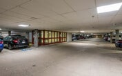 This indoor parking garage is a well-organized, single-level facility designed to accommodate a substantial number of vehicles. The structure features a dropped panel ceiling with evenly distributed fluorescent lighting that illuminates the entire floor, ensuring clear visibility throughout. Wide drive lanes are clearly delineated with painted markings, and individual parking stalls are numbered for easy navigation. Designated accessible parking spaces, marked with the standard accessibility symbol, are conveniently positioned near key access points. A prominent enclosed structure at the center of the facility features a distinctive red-framed glass facade, housing what appears to be a lobby or elevator core, providing a clear interior connection point for occupants. Concrete support columns are spaced throughout the floor, and the perimeter walls are finished in a neutral masonry tone. A dedicated bicycle storage area is visible along one wall, equipped with mounted racks to accommodate cyclists. Stop signage and directional markings are placed at key intersections within the drive lanes to facilitate safe and efficient traffic flow. The overall layout is functional and efficiently designed, prioritizing ease of access, safety, and organization for both drivers and pedestrians navigating the facility.