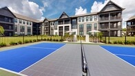 Located at 20 Graf Dairy Drive in Mobile, Alabama, this outdoor pickleball court is a standout recreational amenity designed for active community living. The court features a bold blue and gray modular tile surface with clearly marked lines and a regulation net, providing an ideal setting for both casual play and competitive matches. The court is enclosed by a black metal fence that defines the space while maintaining open sightlines to the surrounding landscape. Lush greenery, ornamental plantings, and manicured mulched beds frame the perimeter, contributing to a well-maintained and visually appealing environment. Adjacent to the court, an outdoor seating area with patio tables and market umbrellas offers a comfortable space for spectators and players to relax. A covered pavilion with a distinctive gabled roofline and dark timber framing is visible nearby, hinting at additional outdoor gathering amenities. The backdrop features a multi-story residential building with craftsman-style architectural details, including peaked gables, board-and-batten siding, and private balconies. The entire setting enjoys abundant natural light under open Alabama skies, creating a bright and inviting atmosphere well-suited for year-round outdoor recreation.