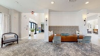 This single-floor community clubhouse blends modern design with warm, inviting interiors throughout its well-appointed spaces. The entry lobby greets visitors with soaring ceilings, large glass doors that draw in abundant natural light, and lush potted greenery, setting an airy and welcoming tone. A stylish lounge area anchors the main gathering space, featuring a tufted high-back banquette, cognac leather seating, and a richly patterned area rug that adds texture and depth.

Adjacent to the lounge, a media room offers a comfortable retreat with a large wall-mounted television framed by a dark, built-in shelving unit with herringbone accent paneling, complemented by a plush neutral sofa and mid-century-inspired armchairs. A separate sitting room provides a quieter setting, furnished with upholstered barrel chairs arranged around side tables beneath large windows overlooking an outdoor terrace.

The fully equipped kitchen is a standout feature, showcasing dark cabinetry, white quartz countertops, a geometric light-teal tile backsplash, stainless steel appliances, and light wood-look flooring. Globe pendant lights and an integrated beverage station add both function and style. The adjacent dining area accommodates multiple tables and chairs beneath wall-mounted displays, with sliding glass doors providing direct outdoor access and filling the room with natural light.