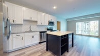 This single-floor unit showcases a thoughtfully designed open layout that connects the kitchen, living area, and bedroom spaces. The kitchen serves as a focal point, featuring white shaker-style cabinetry with matte black hardware, a classic subway tile backsplash, and granite countertops. A full suite of stainless steel appliances includes a refrigerator with ice maker, electric range, over-the-range microwave, and dishwasher. A freestanding island with a light countertop and open shelving below provides additional workspace and storage, finished in a bold contrasting color that adds visual interest to the space.

Light wood-look flooring runs throughout the main living areas, contributing to a clean and contemporary aesthetic. The bedroom offers a comfortable, carpeted floor surface, a ceiling fan, and a window that allows natural light to filter in. The bathroom is finished with a granite-topped vanity with white cabinetry, a tub-shower combination, and matching wood-look tile flooring. A full-size washer and dryer are also included within the unit. Recessed lighting throughout keeps the overall atmosphere bright and welcoming.