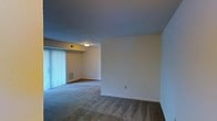 This single-floor residential space offers a straightforward and functional layout designed for comfortable everyday living. The main living area features wall-to-wall carpeting that adds warmth underfoot, complemented by clean, neutral-toned walls that create a bright and versatile backdrop for any furnishing style. A large sliding glass door fitted with vertical blinds serves as a prominent architectural feature, allowing natural light to filter into the room while providing access to an outdoor area. The ceiling is smooth and finished with a flush-mounted light fixture, keeping the space feeling uncluttered and open. A wall-mounted HVAC vent ensures year-round climate control. Transitioning from the carpeted living area, an adjacent room features a contrasting hardwood-style flooring, offering a subtle yet effective delineation of functional zones within the unit. The overall ambiance of the space is clean, neutral, and move-in ready, providing a blank canvas that accommodates a wide range of interior design preferences. The open connection between the two rooms enhances the sense of flow and spaciousness throughout the single-level layout.