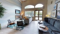 This single-floor communal lounge is designed to impress with its soaring ceilings and open, airy atmosphere. The focal point of the space is a sleek, wall-mounted fireplace framed by a striking floor-to-ceiling marble-effect accent wall, which adds a dramatic and sophisticated touch. A flat-screen television is mounted above the fireplace, making this corner ideal for relaxed viewing. The seating area is thoughtfully arranged with a contemporary gray sectional sofa adorned with vibrant teal, mustard, and patterned throw pillows, complemented by woven accent chairs and low-profile coffee tables that rest atop a soft, ocean-inspired area rug. A second seating cluster with a navy sofa provides additional comfort and flexibility. Large arched transom windows and glass-panel doors flood the interior with natural light while offering views of the surrounding greenery and outdoor pool. A ceiling fan with a wood-toned finish adds to the comfort of the space. Light wood-look flooring runs throughout, tying the design together with a clean, modern aesthetic. The overall ambiance is welcoming and sophisticated, blending warm and cool tones for a balanced, resort-inspired feel.