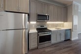 Located at 119 Searing Avenue in Mineola, NY, this thoughtfully designed single-floor residence offers a clean, modern aesthetic throughout its well-appointed spaces. The kitchen serves as a standout feature, showcasing floor-to-ceiling light wood cabinetry with sleek chrome hardware, pristine white quartz countertops, and a subtle tile backsplash with integrated under-cabinet lighting. A full suite of stainless steel appliances — including a gas range, over-the-range microwave, refrigerator, and dishwasher — rounds out this highly functional culinary space. Rich wood-look flooring flows through the kitchen, adding warmth and continuity. The bathroom is finished to an equally high standard, featuring a marble-topped vanity with an under-mount sink, a large backlit mirror, wood-look tile flooring, and a glass-enclosed shower stall with subway tile surround. The bedroom offers a comfortable, carpeted retreat complete with crown molding, recessed lighting, a ceiling fan, and a window with natural light. A built-in closet provides convenient storage. The overall floor plan, as seen from an aerial perspective, offers a compact yet efficient layout with distinct living, kitchen, and sleeping areas seamlessly connected.