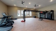 This fitness center is a comprehensively outfitted workout facility designed to support a wide range of exercise disciplines. The space is laid out across a single floor and is divided into distinct functional zones, each dedicated to a different style of training.

The cardio area features a lineup of True-brand treadmills, elliptical machines, and a stationary bike, all positioned in front of a row of windows that allow natural light to fill the room and offer views of the surrounding exterior. Recessed lighting and ceiling fans overhead keep the environment comfortable and well-lit.

Adjacent to the cardio zone is a dedicated functional training area centered around a freestanding multi-use rig. The rig supports suspension trainers, resistance bands, and gymnastics rings, providing versatile options for bodyweight and resistance-based workouts. Durable wood-look flooring runs throughout, offering both resilience and a clean aesthetic.

The strength training section houses a well-stocked dumbbell rack alongside medicine balls and kettlebells, alongside a weight bench and cable machine systems for targeted resistance training. A heavy punching bag adds a combat fitness option to the mix.

Neutral-toned walls and consistent recessed lighting create a clean, modern ambiance throughout. The open floor plan ensures ample room for movement, stretching, and group workouts, making this fitness center a versatile and inviting space for all fitness levels.