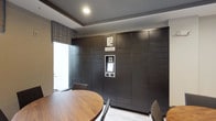 This package concierge room is designed to streamline parcel management with a practical yet polished aesthetic. The focal point of the space is a floor-to-ceiling bank of dark, matte-finished smart lockers branded with the "Package Concierge" system, which spans nearly the entire back wall. The automated locker system is equipped with a central electronic touchscreen interface, allowing residents or occupants to securely retrieve deliveries at any time. The lockers are organized in a grid pattern, offering numerous compartments of varying sizes to accommodate packages of different dimensions. The room features light tile flooring and a tray ceiling with recessed lighting, contributing to a clean and well-lit environment. A large window dressed with striped Roman shades allows natural light to filter into the space, enhancing the brightness of the room. A round wooden table with upholstered chairs is positioned in the foreground, providing a surface for managing parcels or paperwork. The overall ambiance is functional and contemporary, reflecting a thoughtfully designed amenity space built for efficient, secure package handling.