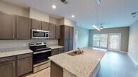 This single-floor residence at 921 Trailwood Dr in Raleigh, NC offers a well-designed layout with contemporary finishes throughout. The kitchen serves as a central highlight, featuring rich dark-stained shaker cabinetry extending to the ceiling for ample storage, light granite countertops, a large island with an integrated sink, and a full suite of stainless steel appliances including a range, over-the-range microwave, and refrigerator. Light wood-style flooring runs through the kitchen and living areas, lending warmth and continuity to the open-concept space. The living area connects seamlessly to the kitchen and includes a ceiling fan and windows overlooking greenery outside.

The bedrooms are carpeted and offer a calm, neutral palette with large double windows that frame views of the surrounding landscape, filling each room with natural light. The bathroom is appointed with a granite-topped vanity, dark cabinetry with chrome hardware, a large wall mirror with overhead lighting, and accessible grab bars. A generously sized walk-in closet adjacent to the bathroom features multi-level wire shelving and hanging rods, providing practical and organized storage. The overall aesthetic is clean, modern, and move-in ready.