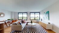This single-floor residence showcases a thoughtfully designed interior with a contemporary aesthetic throughout. Warm hardwood flooring flows seamlessly across the living spaces, creating a cohesive and inviting atmosphere.

The living room is a standout space, featuring multiple large, black-framed windows that frame expansive outdoor views and flood the room with natural light. A bold geometric-patterned area rug anchors the seating arrangement, complemented by a round white coffee table and a rustic wood media console.

The kitchen is well-equipped and modern, featuring two-tone cabinetry with white upper cabinets and warm wood lower cabinets, stainless steel appliances, and a generous central island with built-in shelving and bar seating beneath pendant lighting. An adjacent dining area accommodates a marble-topped table with comfortable seating.

The bedroom is generously proportioned, offering hardwood floors, ample closet space, and a window with outdoor views. Both bathrooms feature floor-to-ceiling wood-look tile walls, soaking tubs with overhead showers, and clean, modern fixtures.

Overall, the space balances a modern design sensibility with warm, natural materials and abundant natural light.