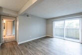 This single-floor residence offers a practical and comfortable layout finished throughout in a clean, neutral color palette. The living area is bright and open, featuring wood-look laminate flooring in a cool gray tone, crisp white baseboards, and a textured ceiling. A large sliding glass door fitted with vertical blinds fills the space with natural light and provides direct outdoor access. Structural beam details add subtle visual interest to the ceiling overhead.

The galley-style kitchen is efficiently arranged with white shaker-style cabinetry lining both walls, offering generous storage above and below. Light-toned laminate flooring, speckled stone-look countertops, and white appliances — including a refrigerator, electric range, and dishwasher — give the kitchen a bright, clean aesthetic.

The bedroom is carpeted for warmth and comfort, with soft gray walls and a window dressed with plantation-style shutters that allow for natural light while maintaining privacy.

The bathroom features a speckled stone-look countertop vanity with a framed mirror, chrome fixtures, and a combined tub and shower. Light wood-look flooring carries through the space, keeping the overall feel cohesive and well-maintained throughout.