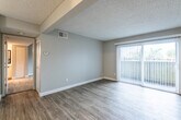 This single-floor residence offers a practical and comfortable layout finished throughout in a clean, neutral color palette. The living area is bright and open, featuring wood-look laminate flooring in a cool gray tone, crisp white baseboards, and a textured ceiling. A large sliding glass door fitted with vertical blinds fills the space with natural light and provides direct outdoor access. Structural beam details add subtle visual interest to the ceiling overhead.

The galley-style kitchen is efficiently arranged with white shaker-style cabinetry lining both walls, offering generous storage above and below. Light-toned laminate flooring, speckled stone-look countertops, and white appliances — including a refrigerator, electric range, and dishwasher — give the kitchen a bright, clean aesthetic.

The bedroom is carpeted for warmth and comfort, with soft gray walls and a window dressed with plantation-style shutters that allow for natural light while maintaining privacy.

The bathroom features a speckled stone-look countertop vanity with a framed mirror, chrome fixtures, and a combined tub and shower. Light wood-look flooring carries through the space, keeping the overall feel cohesive and well-maintained throughout.