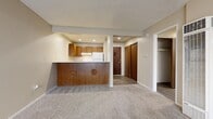 This single-floor, one-bedroom, one-bathroom unit is thoughtfully laid out to maximize its compact footprint. The open-concept kitchen and living area forms the heart of the home, where warm wood-toned cabinetry with decorative panel detailing anchors the kitchen. Light-toned countertops provide ample prep space, and white appliances — including an electric range, dishwasher, and refrigerator — offer full functionality. A peninsula-style counter separates the kitchen from the carpeted living area, which is bathed in natural light through a large sliding glass door fitted with vertical blinds that overlooks an outdoor space. The textured ceiling adds a classic touch throughout the main living areas. The bedroom is a comfortable, carpeted retreat featuring a wall-mounted HVAC unit and a sliding wood-panel closet that provides convenient storage. The bathroom is clean and practical, featuring a wood-vanity with integrated sink, a large wall mirror illuminated by a strip light fixture, and wood-look tile flooring that carries a light, neutral tone. Neutral wall colors and consistent warm wood accents throughout the unit create a cohesive and inviting atmosphere.