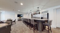 Located at 100 Frankfort Square in Columbus, Ohio, this single-floor community amenity space is thoughtfully designed to offer residents a refined and versatile gathering environment. The main lounge and dining area is anchored by a generously sized kitchen island topped with granite countertops and lined with upholstered bar stools featuring nailhead trim detailing. Stainless steel appliances, including a large refrigerator and wall ovens, are complemented by a bold range hood and geometric pendant lighting that lend the kitchen a contemporary, upscale character. Throughout the open dining area, multiple round wooden tables are surrounded by grey upholstered armchairs with nailhead accents, arranged to encourage both intimate gatherings and larger social events. Decorative white columns with detailed molding add architectural interest, while curated wall art — including geometric wood-panel pieces and a vivid cityscape painting — contribute to the space's polished aesthetic. A wall-mounted flat-screen TV enhances the lounge area. Adjacent to the main gathering space, a dedicated media room offers a more intimate atmosphere, featuring plush cognac leather recliners, mounted surround sound speakers, a large flat-screen TV, and a curated selection of media for viewing. Warm sconce lighting and darker tones in this room create an immersive, cinema-like experience.
