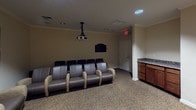 This single-level space is purpose-built for both cinematic and musical experiences, combining the functionality of a screening room with a versatile gathering area. The room is anchored by a large wall-mounted projection screen paired with a ceiling-mounted projector, delivering a true theater atmosphere. Rows of plush, gray leather theater-style chairs with upholstered armrests are arranged to face the screen, providing comfortable seating for a group audience.

Crown molding lines the ceiling throughout, adding a refined architectural detail, while recessed lighting and patterned carpeting contribute to the room's warm, inviting ambiance. The walls are finished in soft neutral tones, enhancing the understated elegance of the space.

Along one side of the room, a built-in wooden cabinet with a granite countertop offers a practical service or refreshment area. A wooden storage console sits near the screen, providing additional functionality. Completing the room's versatile character is a full electronic organ with pedals and candle holders, positioned along the opposite wall — suggesting the space serves ceremonial or multi-use purposes as well. An exit door provides convenient access, and the overall layout is thoughtfully organized to accommodate both intimate gatherings and larger group events.
